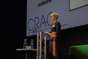 A woman stands at a podium presenting to an audience with a giant screen behind her.