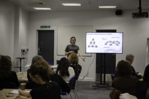 A woman stands at the front of a seminar room and leads a workshop; there is a screen behind her to show her presentation and people are sat at tables listening.