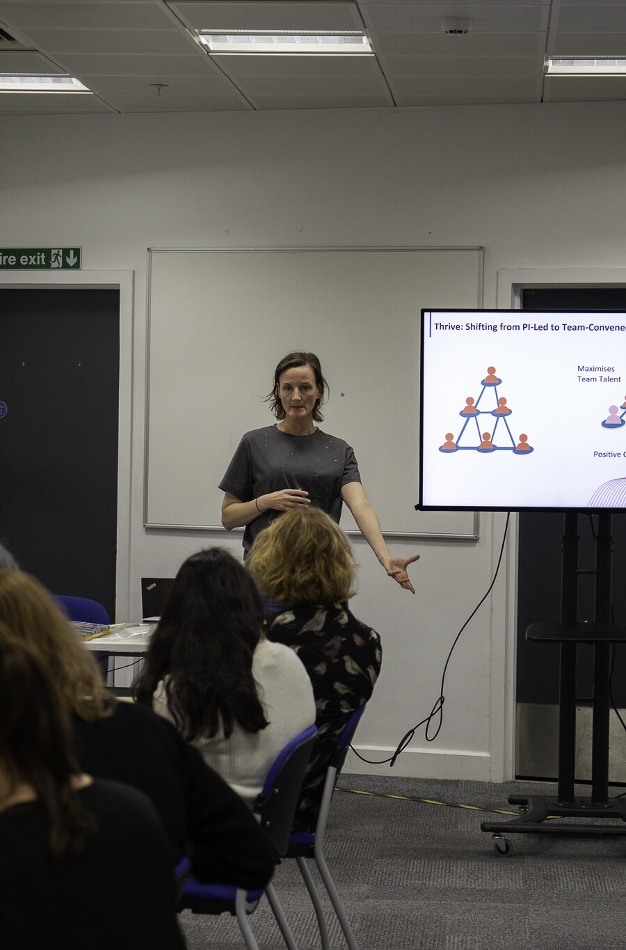 A woman stands at the front of a seminar room and leads a workshop; there is a screen behind her to show her presentation and people are sat at tables listening.