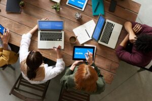 People sitting around a table, seen from an aerial view. There are laptops on the desk with them. Photograph from Pexels. 