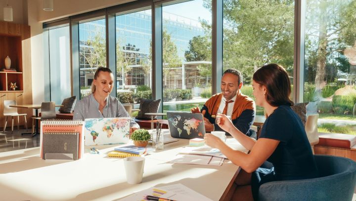 Three people sit around a table with laptops and other stationery in front of them. Behind them is a big glass window that looks out onto tress and sky. Photograph from Pexels by Kindel Media.