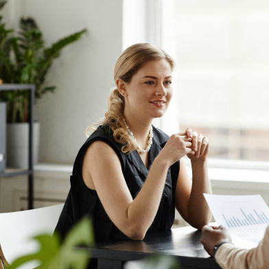 A woman sat at a desk with her elbows propped on the desk, listening to another person. Windows are in the background.