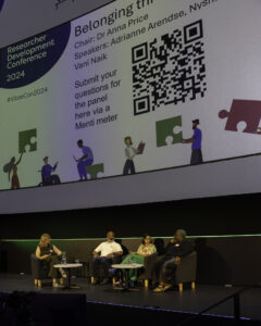 Four people sit in chairs on a stage at a panel discussion with a giant screen behind them. Photograph from VitaeCon2024. 