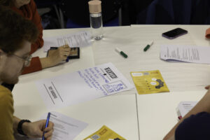 A view of a table with paper and pens strewn across it, with the sides of peoples' faces just out of the frame.