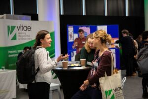 A group of women stand talking in a conference centre with the Vitae logo on a banner behind them.