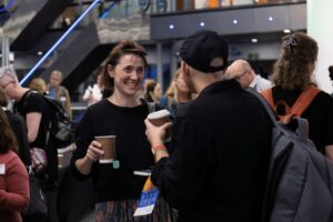 A woman talks with a man, who has his back to the camera. Both are holding coffee cups.