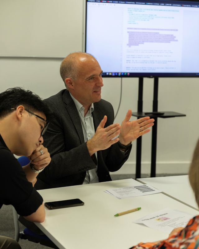 A picture of a man sat at a table with other people around him; in the background is a presentation screen.