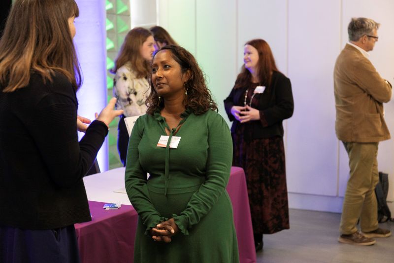 A woman wearing a green dress stands and listens to another woman talking, there are various other people talking to each other in the background.