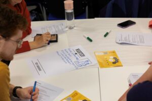 A table with pens and paper strewn across it; people are gathered around the table discussing and problem solving.