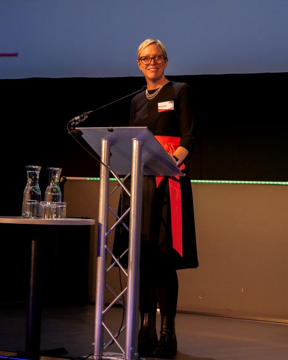 A woman wearing a black dress with a red sash stands at a lectern in front of a screen to give a lecture.