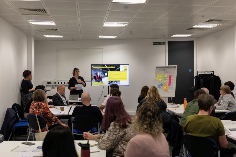 A room full of people in a seminar workshop, looking at a woman presenting in front of a presentation screen.