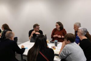 A group of people in a white walled room sat around a table discussing in a workshop.