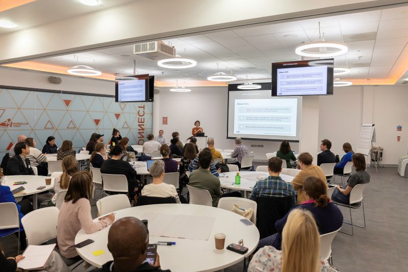 A seminar room full of people sat at round white tables listening to a presenter during a workshop.